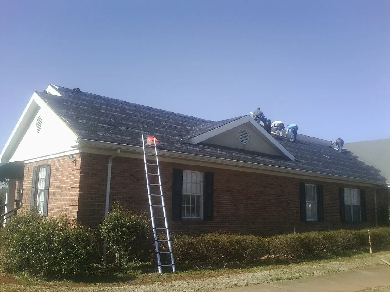 Crew tearing shingles off a brick ranch roof with a ladder set — Metal Roof Repair work by our crew in Starkville