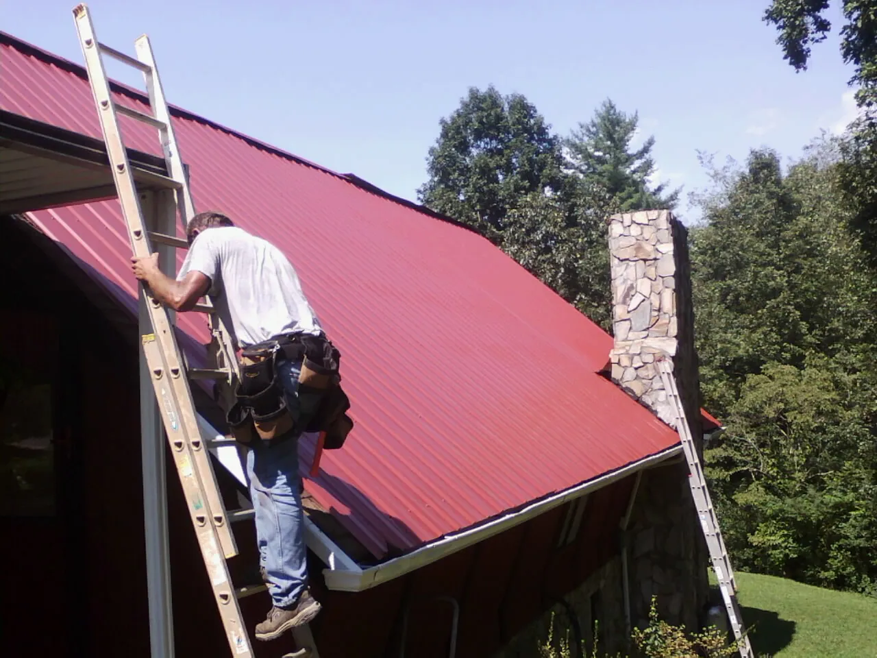 Roofer on a ladder beside a red metal roof and stone chimney — Soffit & Fascia Repair by Roofing Brothers in Starkville